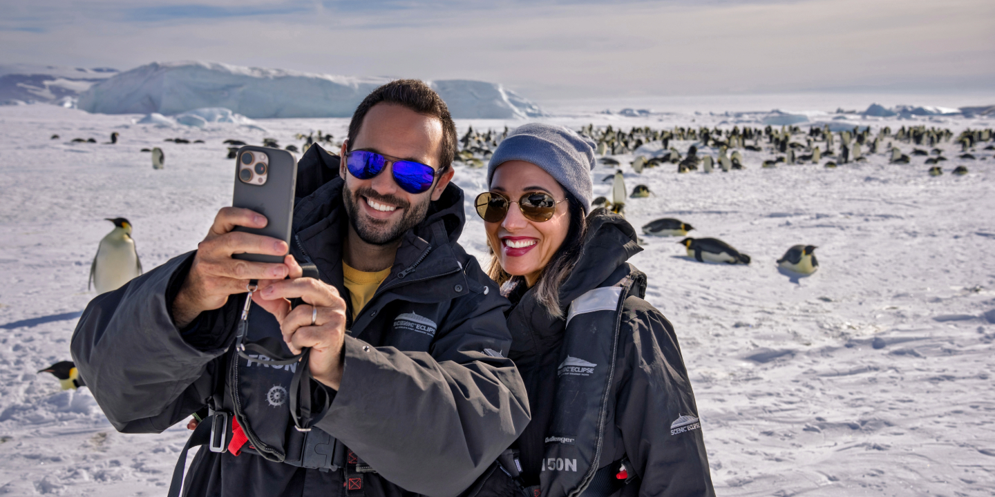 A man and woman capturing a photo of themselves on an iPhone on snow hill island, stood in front of a penguin colony.