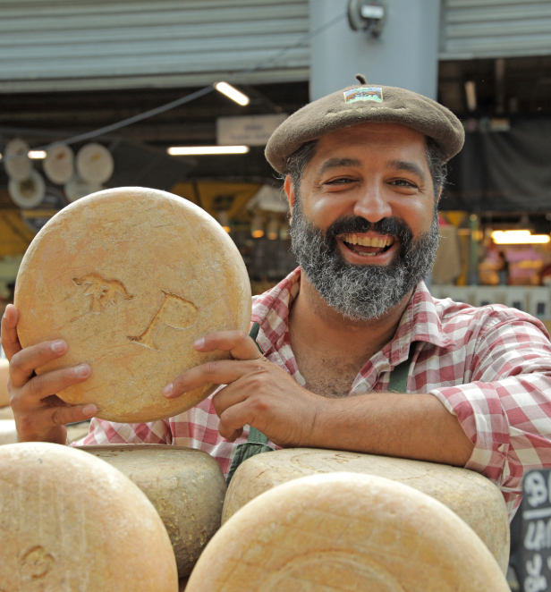 A cheesemonger pictured holding a wheel of cheese behind a stall, dressed in a check shirt and brown hat.