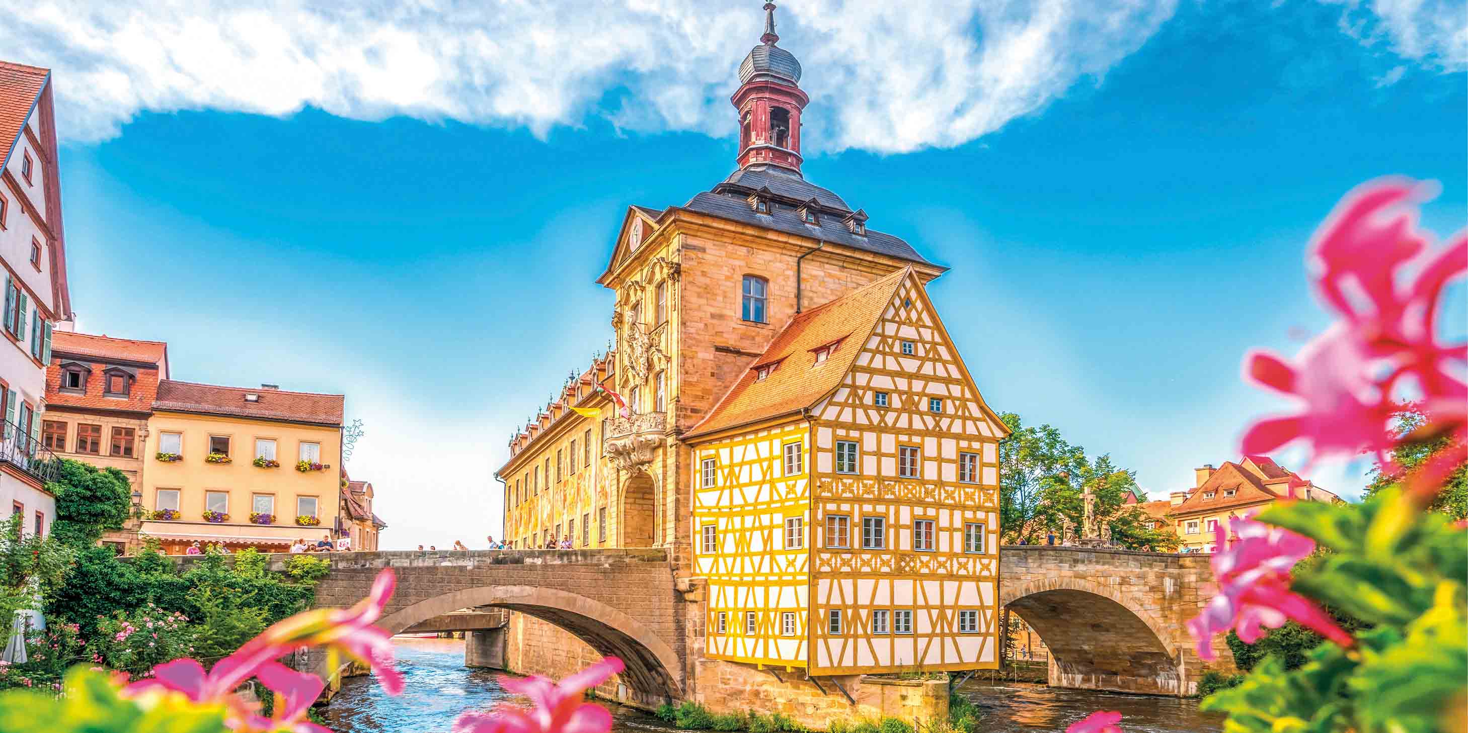 A spring image of Bamberg Bridge, Germany. The sky is blue and pink flowers can be seen in the foreground.