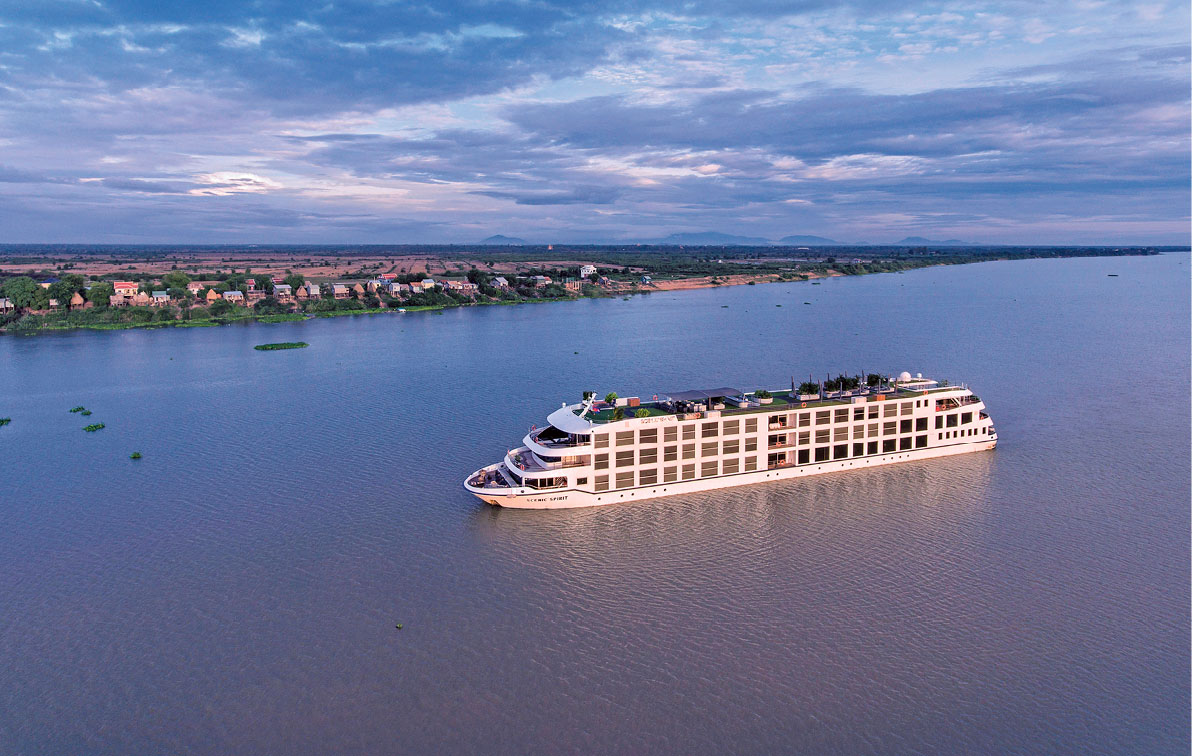 White cruise ship sailing on the Mekong river at sunset with cloudy blue skies and the shoreline with small houses in the distance 