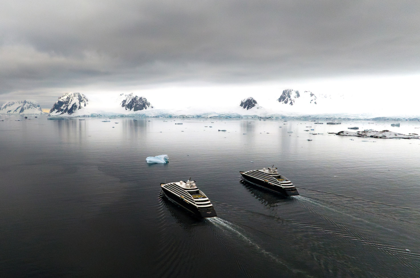 Scenic Eclipse I and II sail away together in Antarctica with black and white icebergs in the background and a grey cloudy sky