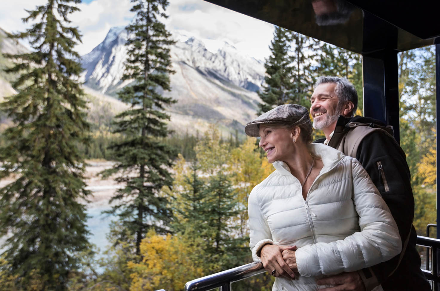 A couple stand smiling looking out of the viewing platform of the Rocky Mountaineer train as trees and mountains roll by