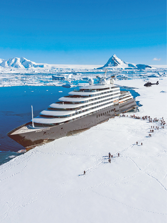 Luxury cruise ship docked on the edge of an ice sheet in Antarctica with groups of people leaving the ship to explore on the ice under sunny blue skies with a helicopter overhead.