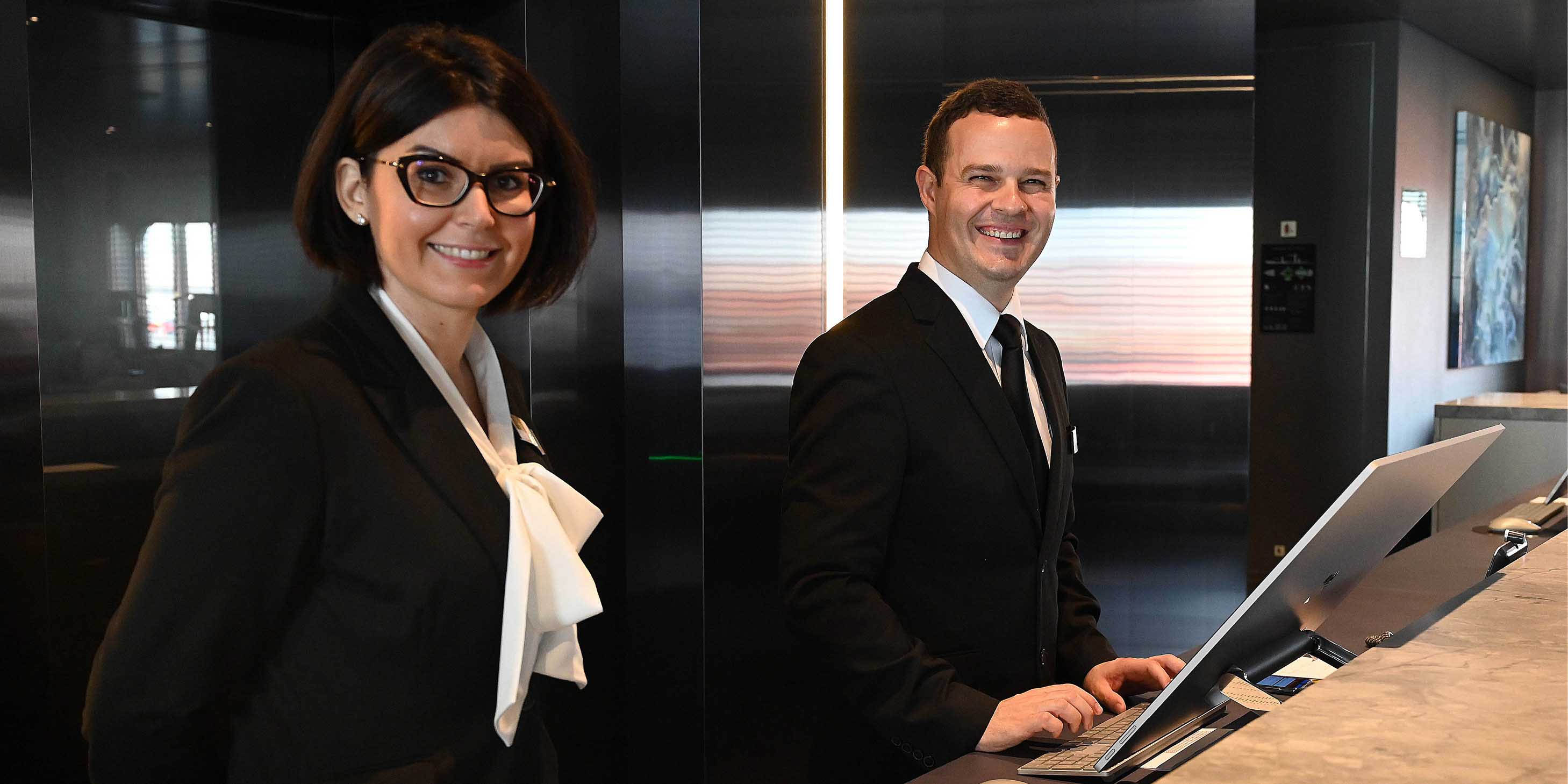 a male and a female member of reception staff on board Scenic Eclipse stand smiling at the desk ready to assist