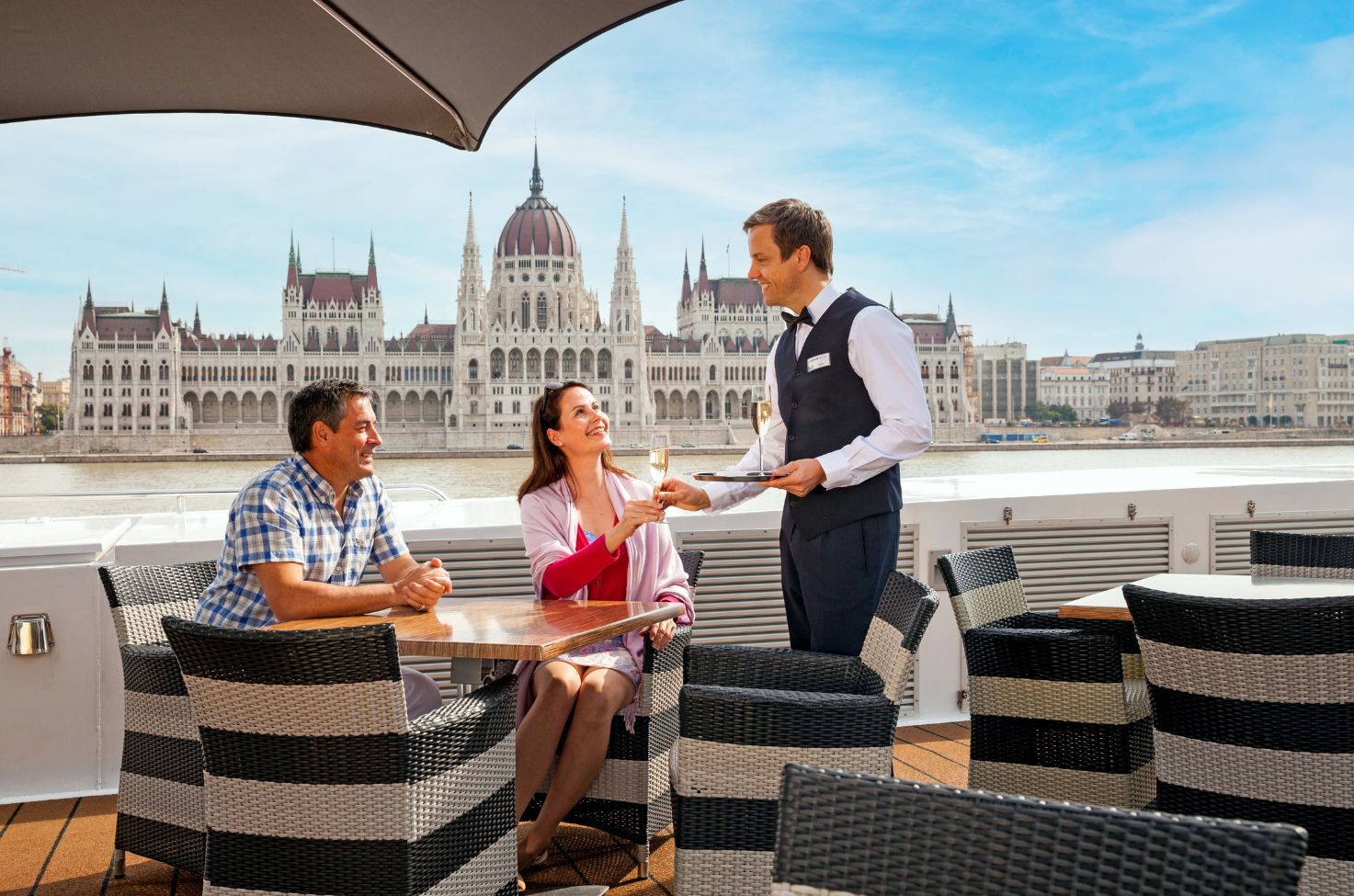 Butler serving a couple at a table on the roof of a river cruise ship with the Budapest Parliament Building in the background
