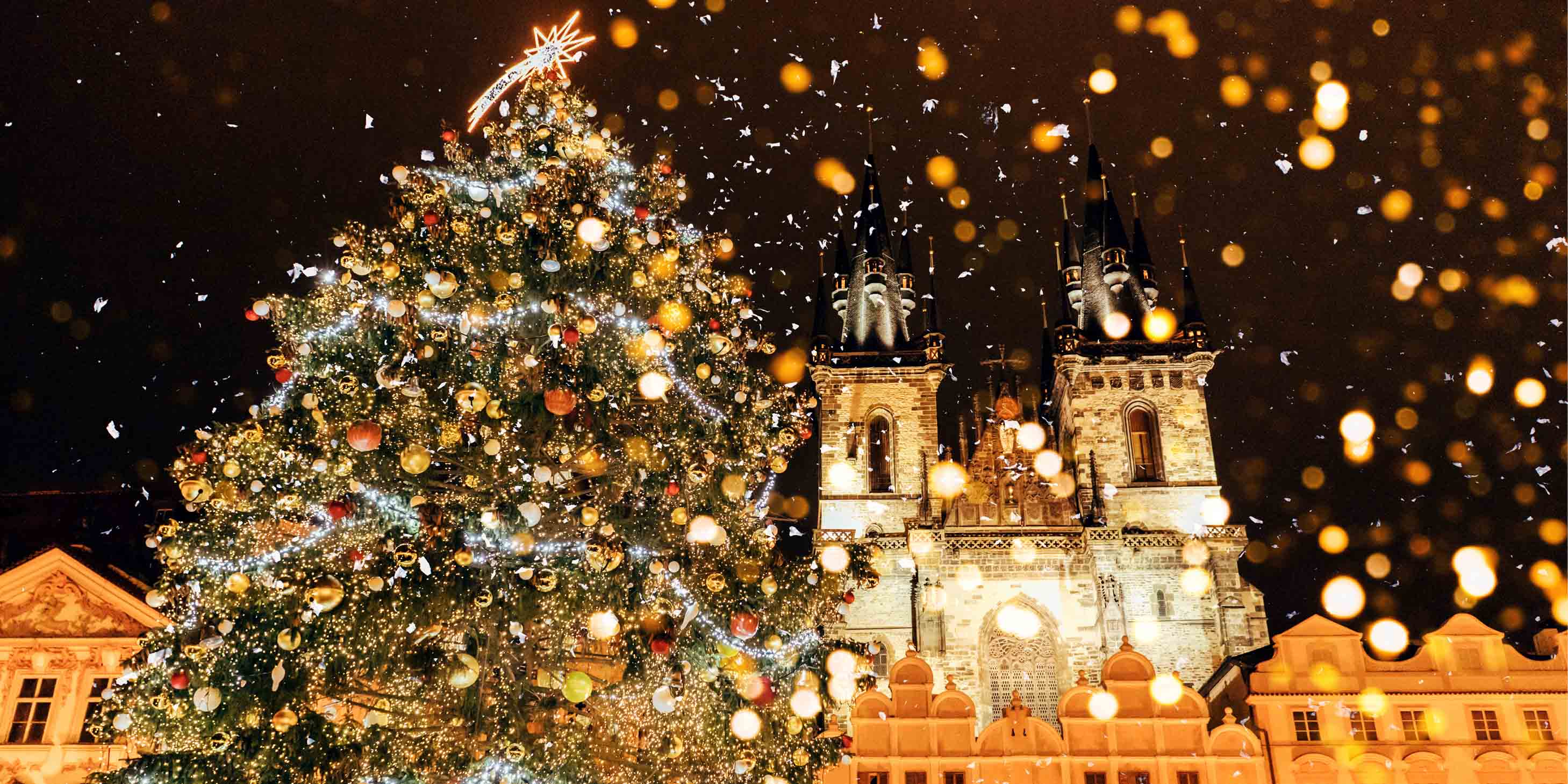 A christmas tree covered with lights, baubles and decorations in front of an illuminated building in Prague.  