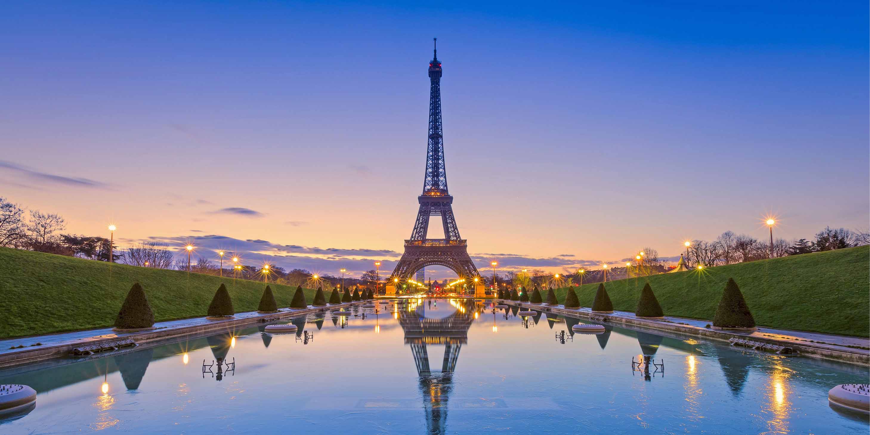 The Eiffel Tower in Paris at dusk with a body of water in front showing the reflection of the tower and lights 