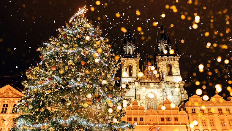 A christmas tree covered with lights, baubles and decorations in front of an illuminated building in Prague.  