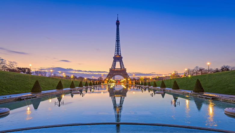 The Eiffel Tower in Paris at dusk with a body of water in front showing the reflection of the tower and lights 