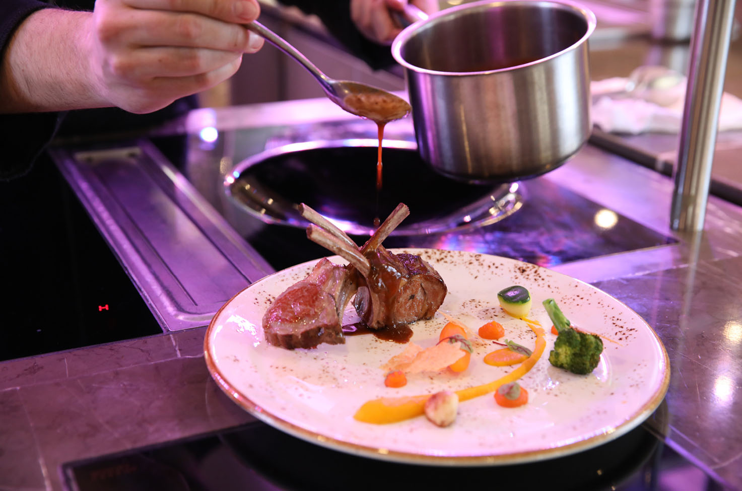 Sauce is drizzled over lamb cutlets presented neatly with various vegetables and other sauces on a white plate