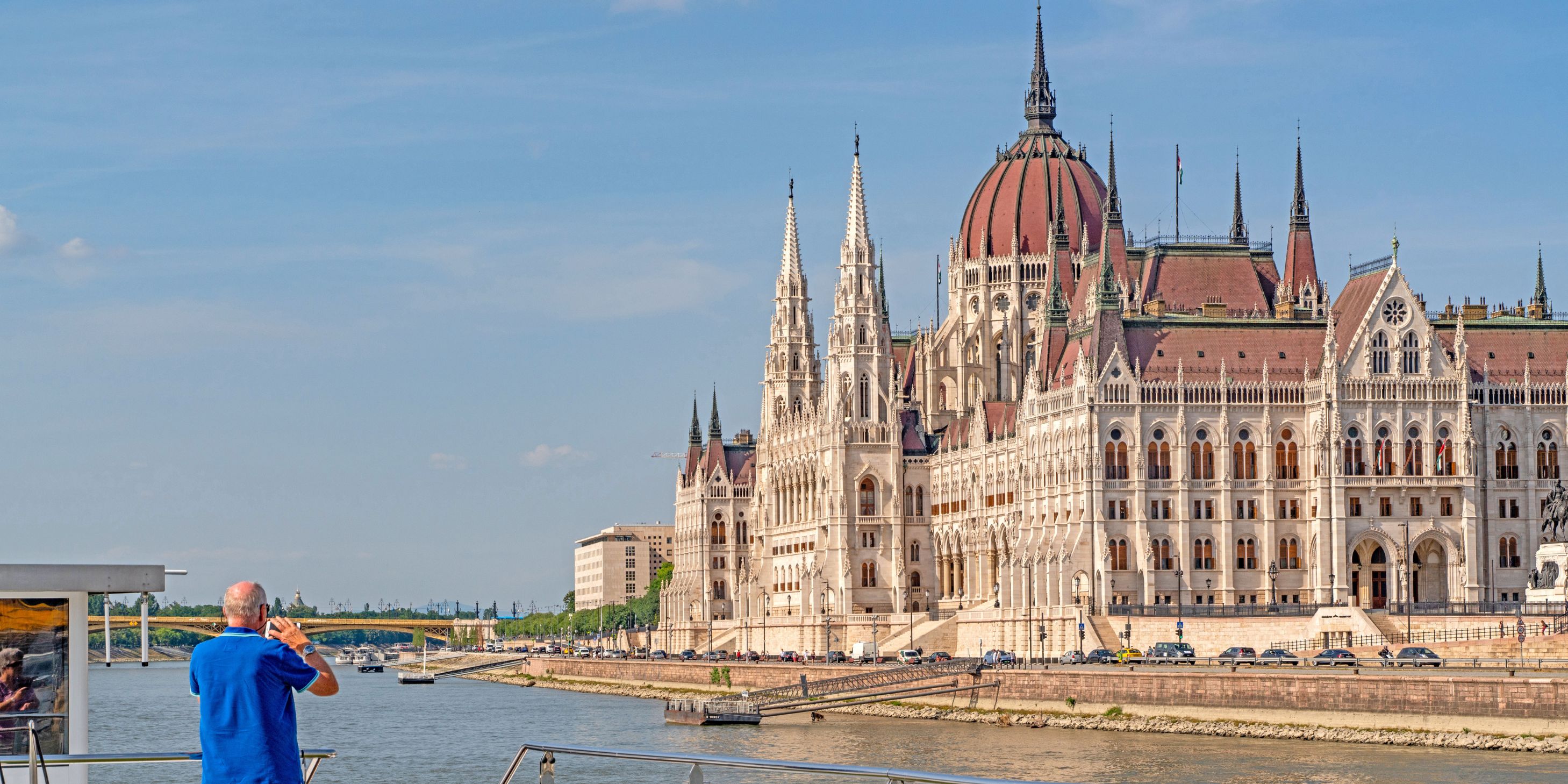 Budapest Parliament Building from the Sun Deck of a Scenic cruise ship on a clear day. A man dressed in a blue t shirt and shorts takes a photo of the view on his smartphone. 