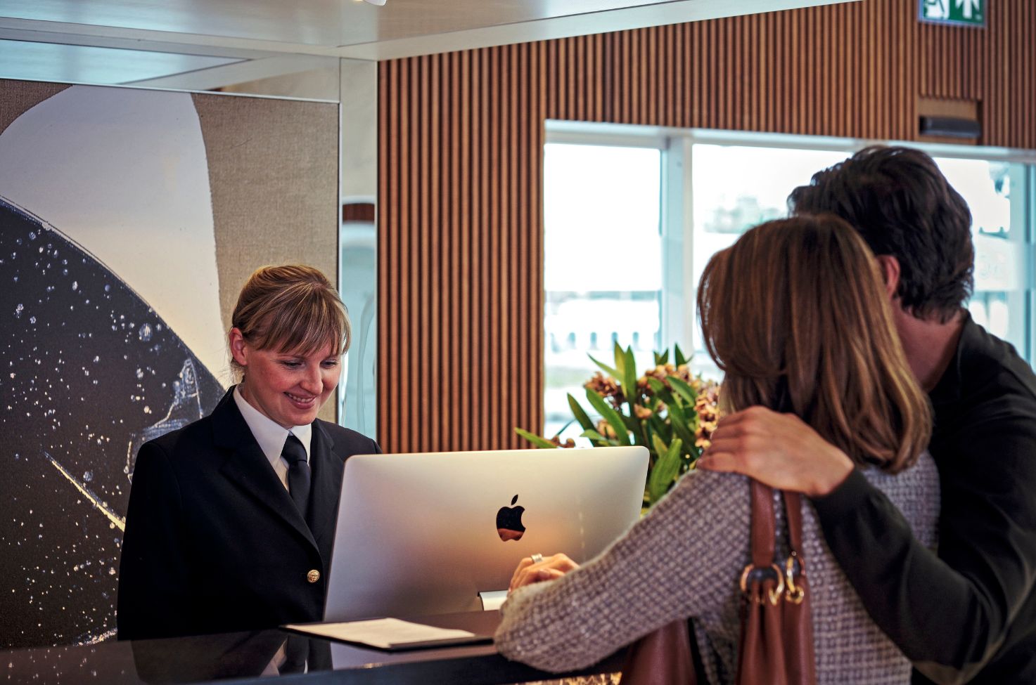 A member of Scenic crew stands at reception smiling at a mac computer as a man and woman wait to be checked in 